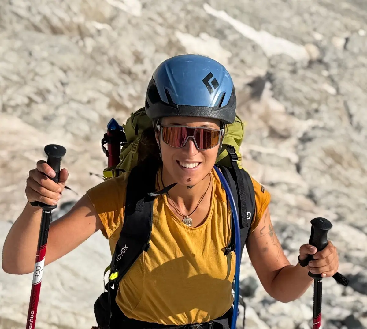 Carmen in a helmet and sunglasses, walking poles in hand, climbing a sunlit snowfield.