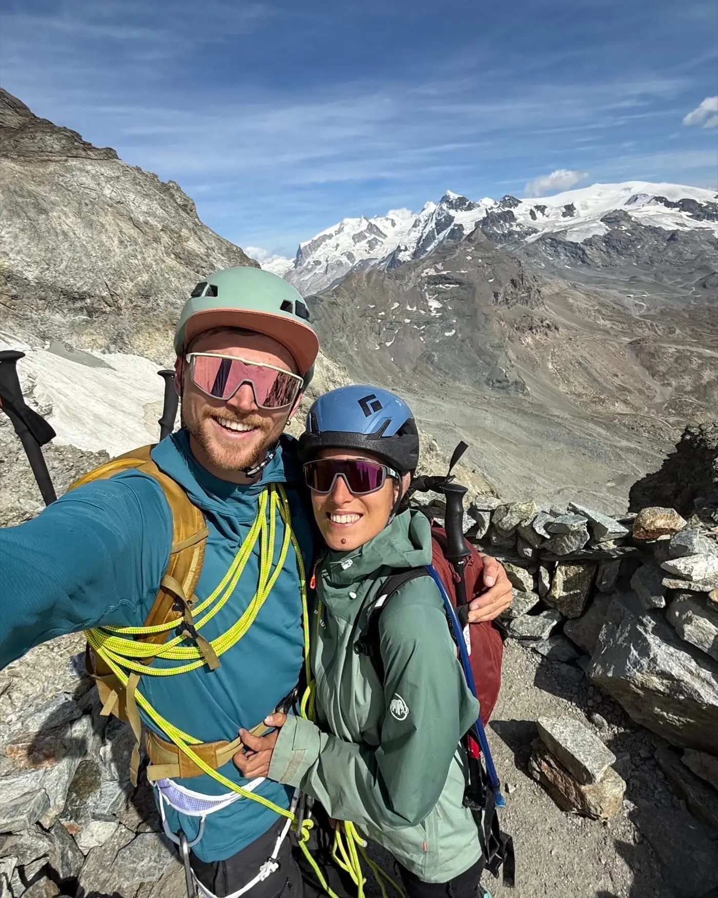 Charlie and Carmen on a rocky ridge in helmets and harnesses, a rope slung across Charlie’s chest, with glaciated peaks behind them.