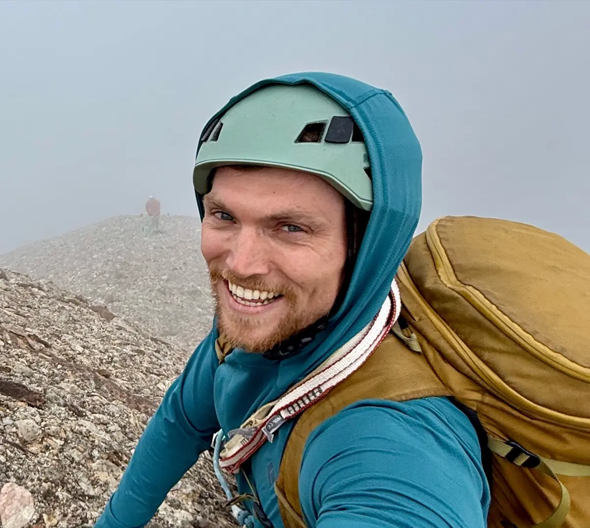 Charlie grinning in a hooded jacket and climbing helmet on a misty ridge, a climbing partner on the skyline behind him.