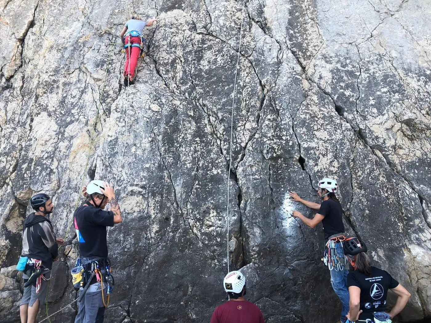 A group in helmets at the foot of a grey limestone crag, watching a climber working the moves high above them.