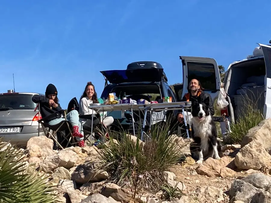 Three people eating at a folding table between a car and a van below a crag, a border collie sitting on the rocks beside them.
