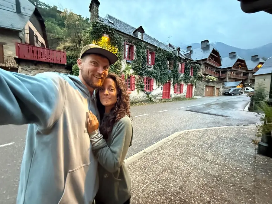Charlie and Carmen in a Pyrenean village at dusk, ivy and red shutters on the stone houses behind them.