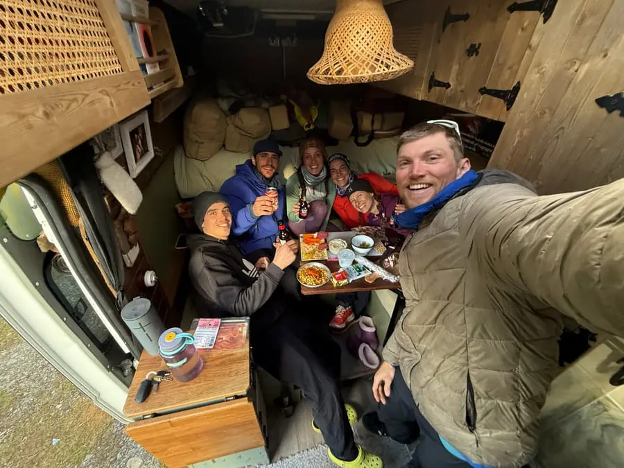 Six people crammed onto the bed in the back of a van, eating together and raising bottles to the camera.