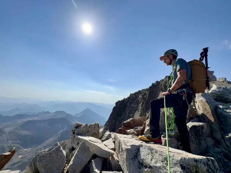 A climber coiling the rope on a granite summit ridge high in the Maladeta massif, range after range of Pyrenean peaks fading into the haze below.