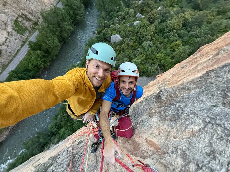 Two climbers at a belay high on a wall, looking down at the camera with the forest far below.