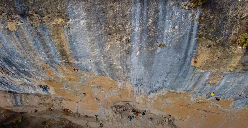 The blue-streaked wall of the Contrafort de Rumbau at Oliana, a climber high on the steep centre of the crag and belayers gathered on the terrace below.
