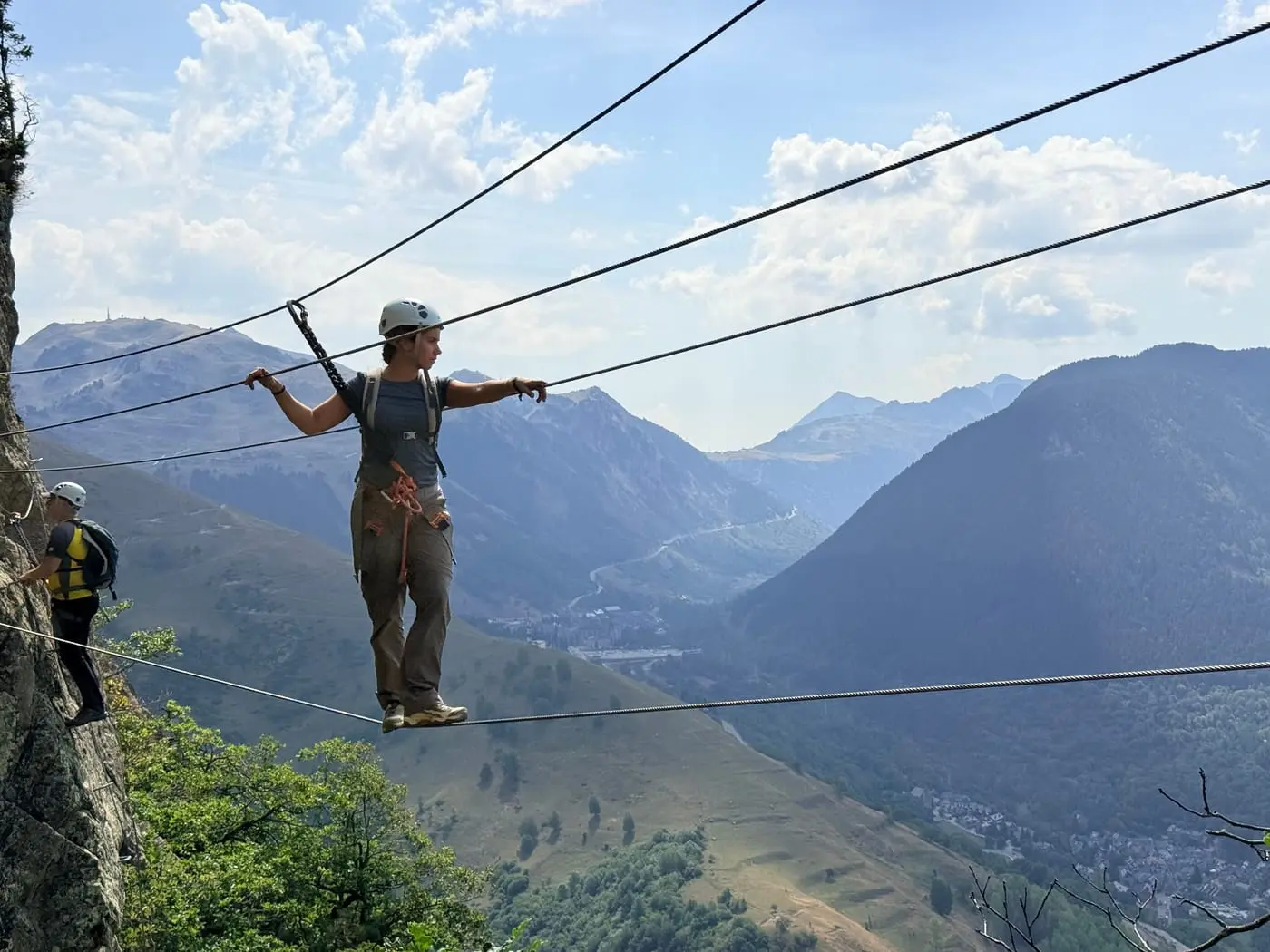 A climber crossing a wire bridge on a via ferrata, arms out on the hand cables, with a green valley far below.